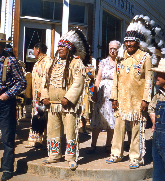 Central figures are two Native American men dressed in native clothing and war bonnets. Background persons include men in western clothing and an older female in a floral dress.